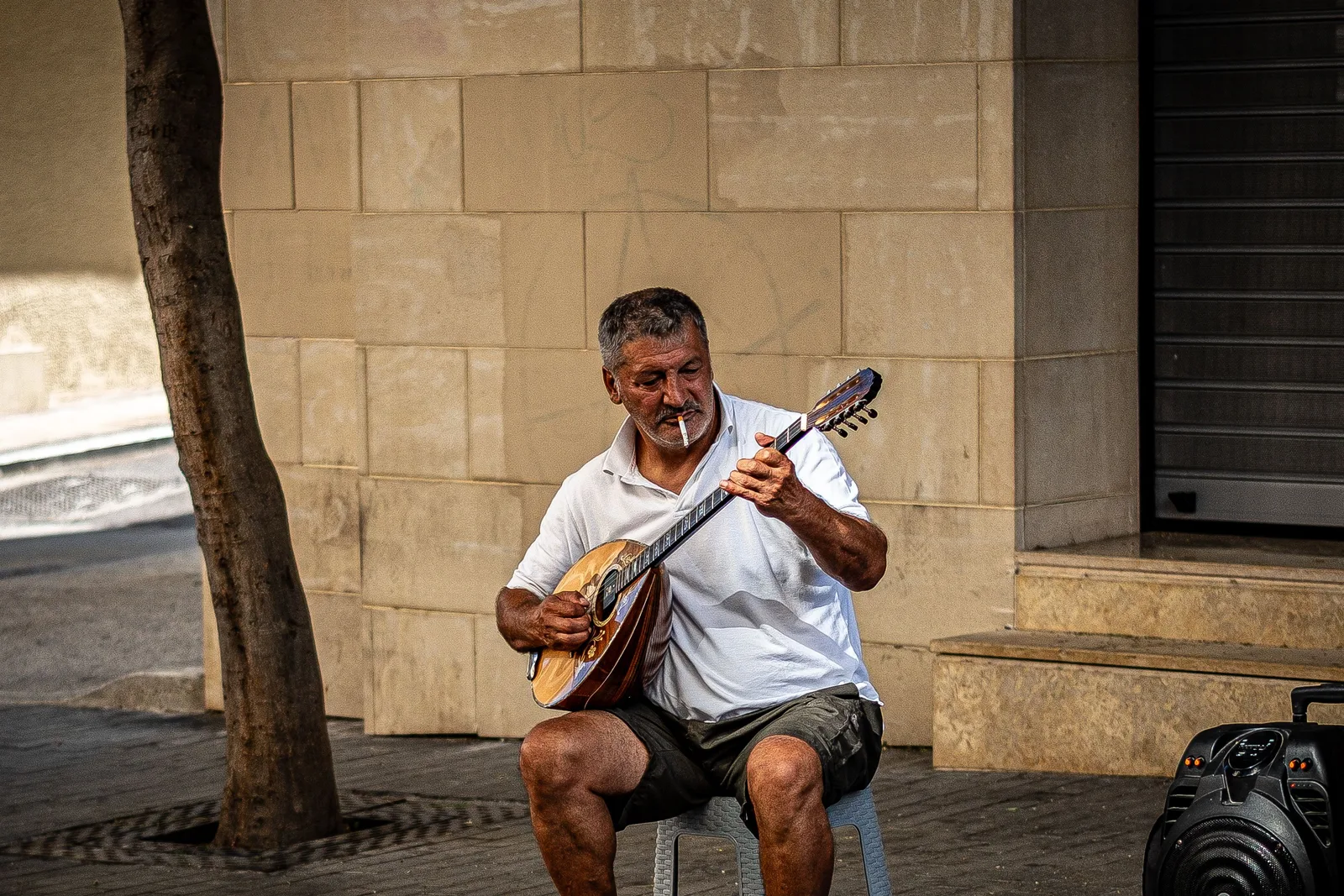 Musicien de rue en Grèce jouant du bouzouki, cigarette aux lèvres, assis devant un mur de pierre