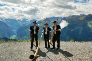 Trois joueurs de cor des Alpes en costume traditionnel suisse tenant leurs longs instruments face a un panorama de montagnes