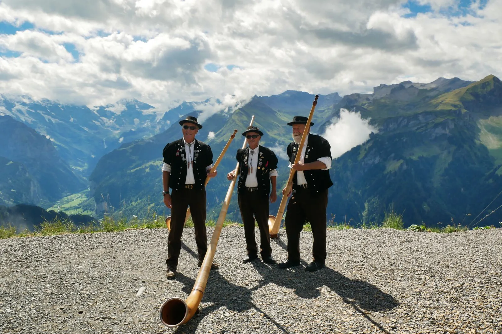 Trois joueurs de cor des Alpes en costume traditionnel suisse tenant leurs longs instruments face a un panorama de montagnes