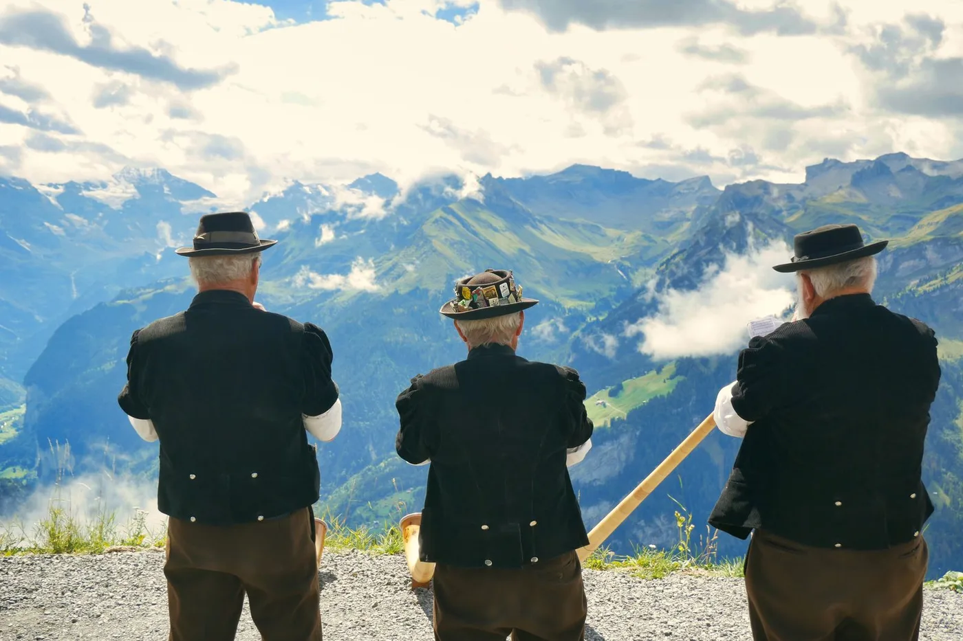 Trois sonneurs de cor des Alpes en costume traditionnel suisse, vus de dos, face a un panorama de montagnes