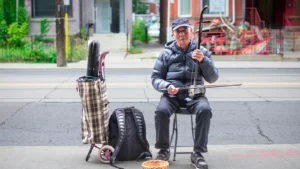 Musicien age jouant de l'erhu assis sur un trottoir en ville, la caisse hexagonale de l'instrument bien visible