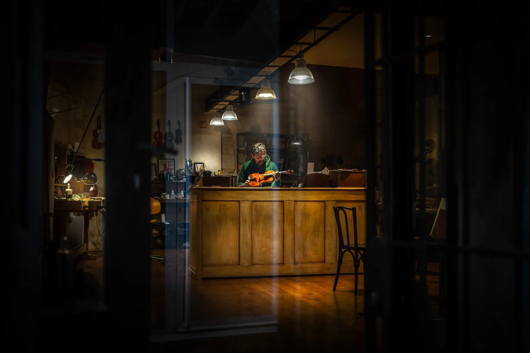 Un luthier examine un violon sous une lampe, vu à travers la vitrine de son atelier, violons suspendus en arrière-plan